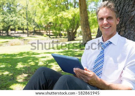 Portrait of happy businessman with digital tablet leaning on tree trunk in park