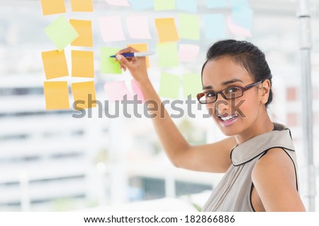 Smiling designer writing on sticky notes on window looking at camera in creative office