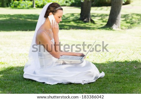 Side view of young bride using laptop and mobile phone while sitting on grass in park