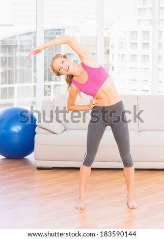 Fit blonde smiling at camera while stretching at home in the living room