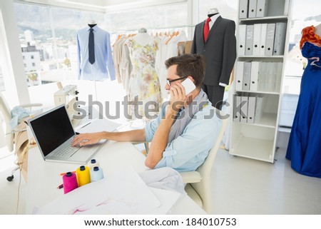 Side view of a young male fashion designer using laptop and cellphone in the studio
