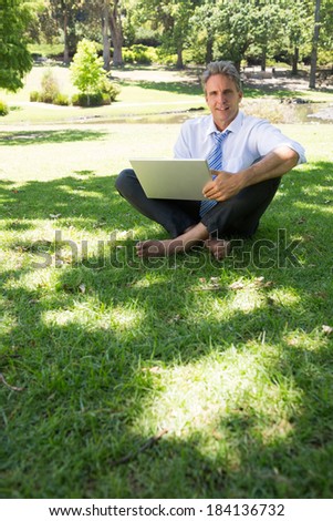 Full length portrait of confident businessman with laptop sitting on grass in park