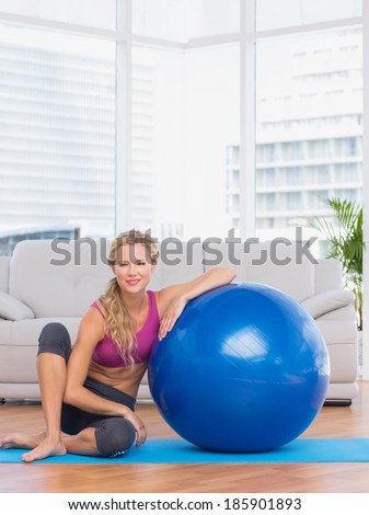 Slim blonde sitting beside exercise ball smiling at camera at home in the living room