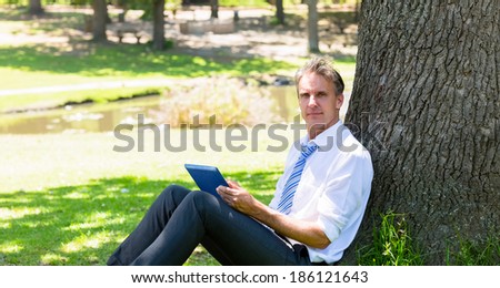 Portrait of mature businessman with tablet computer leaning on tree trunk in park