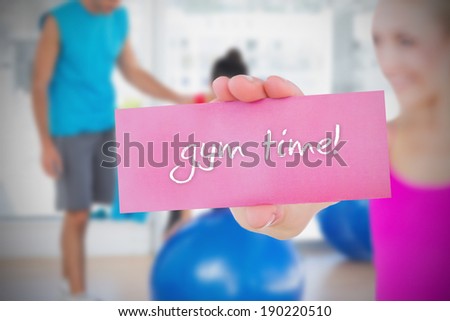 Fit blonde holding card saying gym time against trainer and client in fitness studio