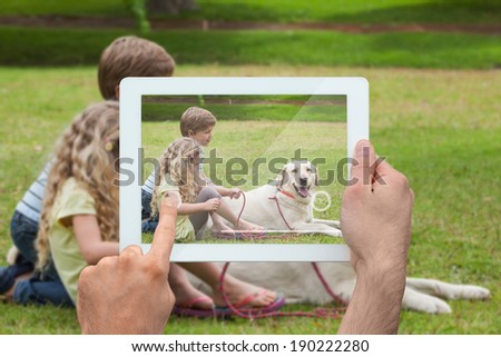 Hand holding tablet pc showing cute siblings sitting with pet dog in park