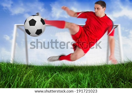 Fit football player jumping and kicking ball against field of grass under blue sky