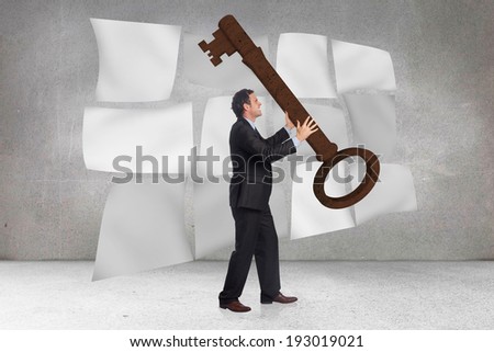 Stressed businessman carrying large key against floating sheets in front of grey wall