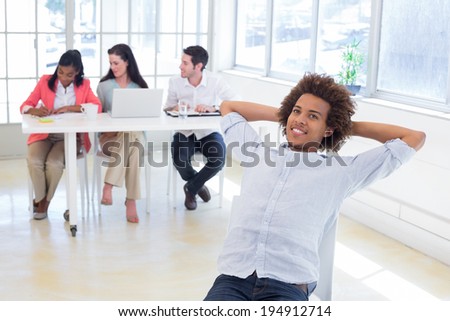 Businessman relaxing on a swivel chair smiling while his coworkers are working behind him