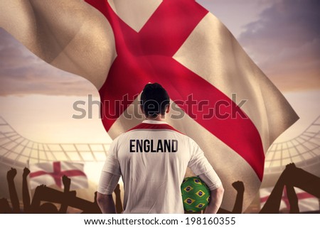 England football player holding ball against large football stadium under cloudy blue sky