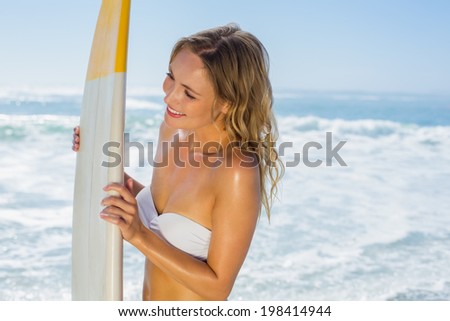 Smiling blonde surfer in white bikini holding her board on the beach on a sunny day
