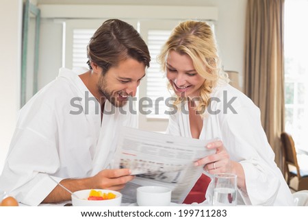 Cute couple in bathrobes having breakfast together reading the newspaper at home in the living room