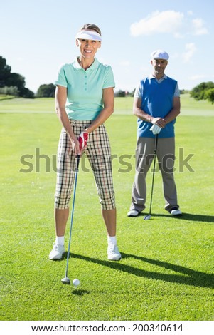Smiling lady golfer teeing off for the day watched by partner on a sunny day at the golf course