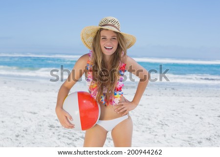 Fit blonde in white bikini and straw hat holding beach ball on a sunny day