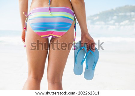Lower half of fit woman holding flip flops on beach on a sunny day