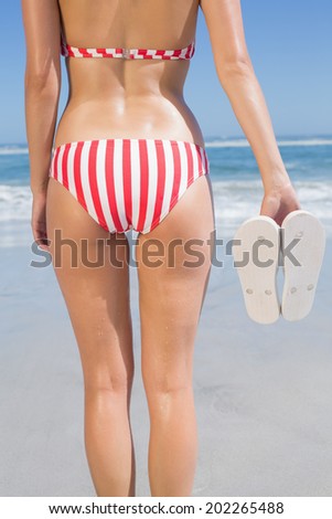 Mid section of fit woman in bikini on the beach holding flip flops on a sunny day