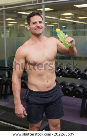 Shirtless bodybuilder drinking sports drink smiling at camera at the gym