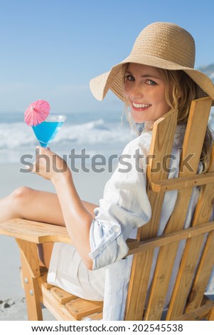 Smiling blonde relaxing in deck chair by the sea holding cocktail on a sunny day