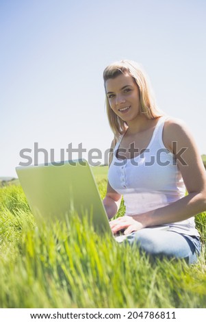 Pretty blonde sitting on grass using her laptop smiling at camera on a sunny day in the countryside