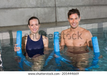 Happy couple holding foam rollers smiling at camera in swimming pool at the leisure centre