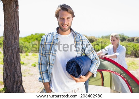 Attractive man smiling at camera while partner pitches tent on a sunny day