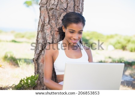Fit woman sitting against tree using laptop on a sunny day in the countryside