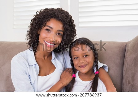 Pretty mother sitting on the couch with her daughter smiling at camera at home in the living room