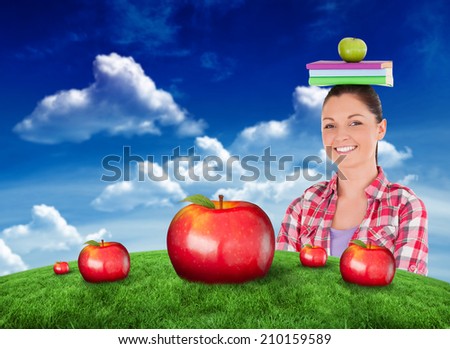 Pretty student holding an apple and books on her head against green field under blue sky