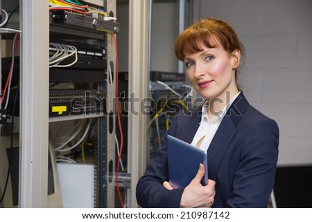 Pretty technician smiling at camera beside open server holding tablet pc in large data center
