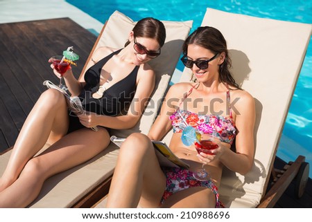 Two beautiful young women with drinks reading books by swimming pool