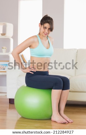 Fit brunette sitting on exercise ball smiling at camera at home in the living room