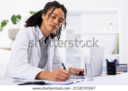 African american mid adult female doctor writing on clipboard while using laptop on desk in hospital. wireless technology, prescription, doctor, medical, healthcare, hospital and medical occupation. Imagine de stoc © 