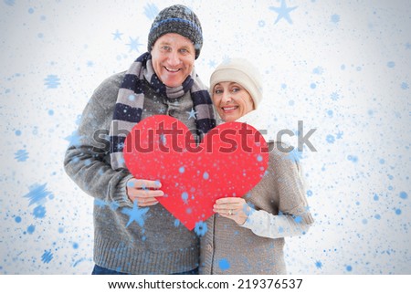 Happy mature couple in winter clothes holding red heart against snow falling