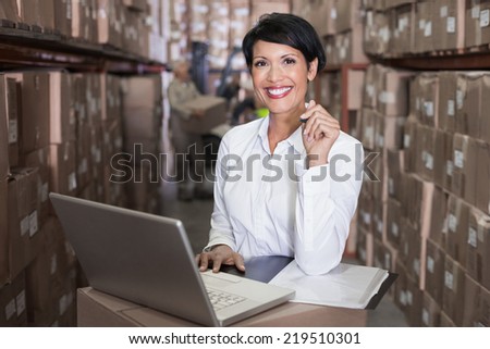 Pretty warehouse manager smiling at camera using laptop in a large warehouse