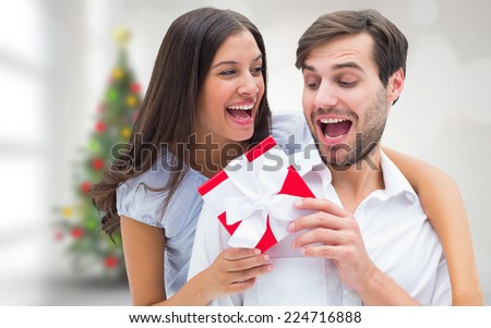 Woman surprising boyfriend with gift against blurry christmas tree in room