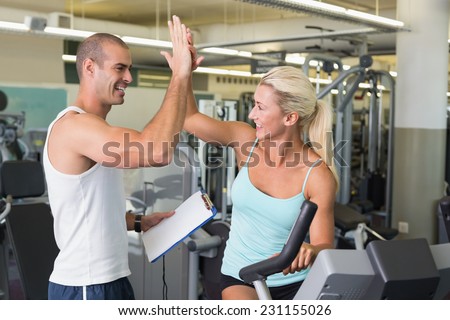 Smiling male trainer giving high five to his client on exercise bike at gym
