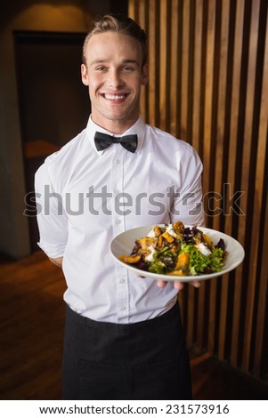 Smiling waiter showing plate of salad to camera in a bar