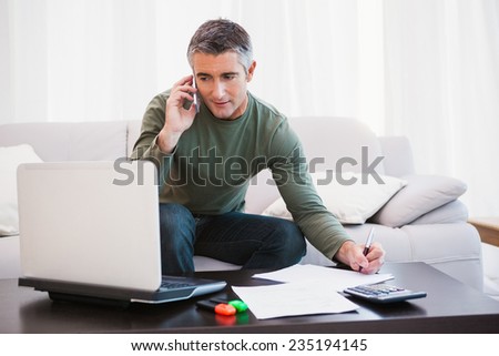 Man with laptop phoning and taking notes at home in the living room