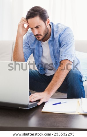 Worried man sitting at table using laptop to pay his bills in the living room
