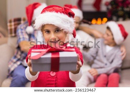 Smiling little girl offering a gift with her parents behind at home in the living room
