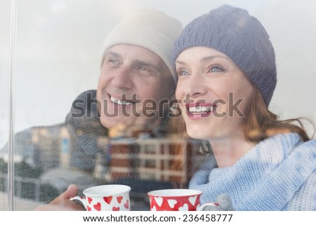 Happy couple in warm clothing holding mugs seen through glass window