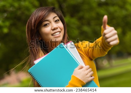 Portrait of female college student with books gesturing thumbs up in the park
