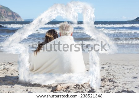 Couple sitting on the beach under blanket looking out to sea against house outline in clouds