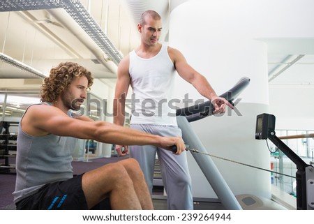 Side view of a male trainer assisting young man on fitness machine at the gym
