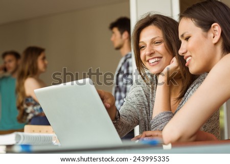 Smiling friends sitting using laptop near classmates at school