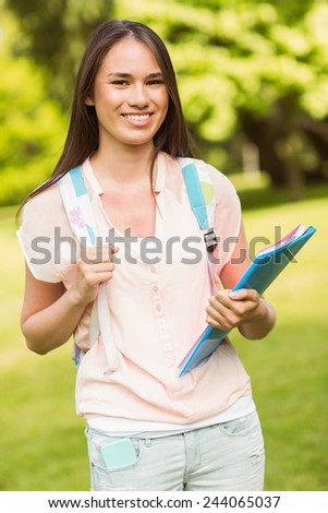 Portrait of a smiling student with a shoulder bag and holding book in park at school