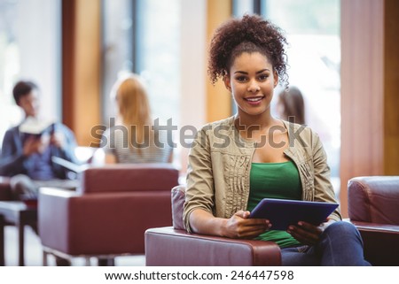 Student sitting on sofa using her tablet pc smiling at camera at the university
