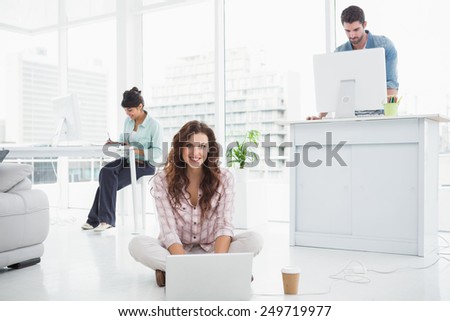 Happy businesswoman sitting on the floor using laptop with colleagues behind her