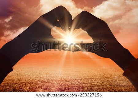 Woman making heart shape with hands against sunrise over field