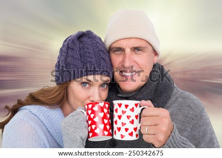 Happy couple in warm clothing holding mugs against room with large window looking on city